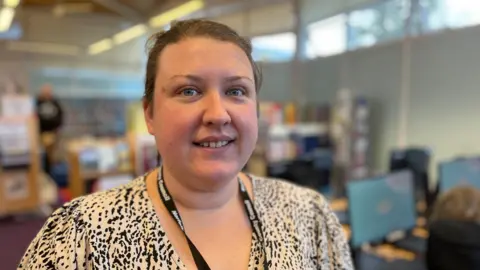 JO THEWLIS/BBC Holli Holmes is wearing a black and white dress and a black lanyard around her neck. She is looking into the camera with a bank of computers in the background.