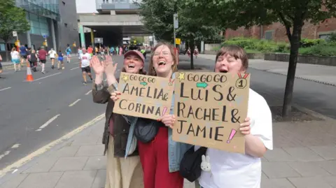 North News Three young women cheering on runners. One is holding a placard made out of a sheet of cardboard saying "Go Go Go Louis & Rachel & Jamie. Another says Go Go Go Amelia & Corina. All are smiling and cheering, with the one without a placard clapping her raised hands. Runners can be seen on the road next to them, John Dobson Street.
