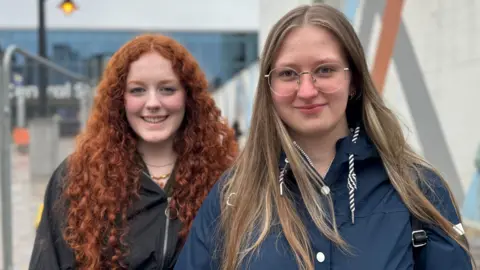 Two girls smiling at the camera. Erain has curly ginger hair and Katie has long blond hair. They are standing outside a train station in Belfast.
