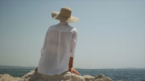 A woman sat in the sun wearing a loose shirt and sun hat