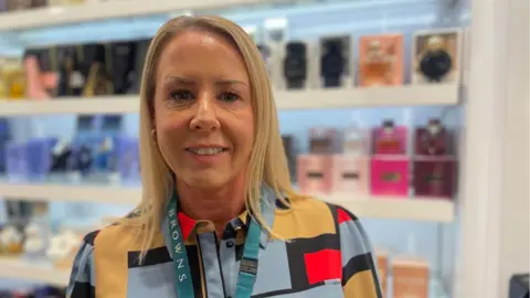 A headshot of Sarah Walker taken in the perfume department at Browns department store. Behind her are multi-coloured perfume bottles in a large, glass-fronted cabinet. Sarah has shoulder length blonde hair, is smiling, and is wearing a shirt dress with a multi-coloured geometric pattern. She has a blue lanyard with Browns written on, round her neck. 