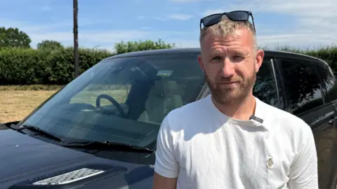Gabby Colenso/BBC Mark Reardon stands wearing a white T-shirt in front of a dark grey 4x4 vehicle looking at the camera. He is squinting slightly because of the sunlight and has sunglasses on the top of his head. The sky behind him is blue with thin, whispy clouds. 