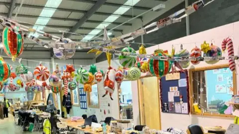 John Devine/BBC Christmas inflatable decorations are strung across the ceiling of an industrial building, with cluttered tables down the middle of the space.