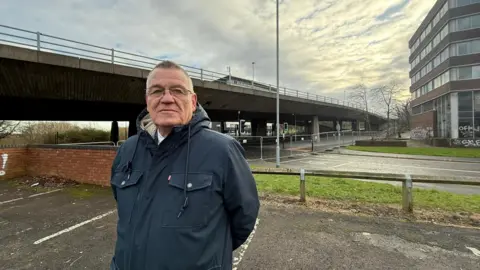 Martin Gannon standing in a car park near the Gateshead flyover. He has short hair and is wearing glasses and a navy coat. The concrete bridge is behind him. Temporary fencing has been put up across the road running under the bridge.