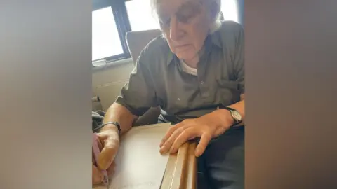 Orchard House Care Home Wally is sitting in a beige chair and is writing a note on a white piece of paper. He has short grey hair and is wearing a white vest with a green shirt over the top.