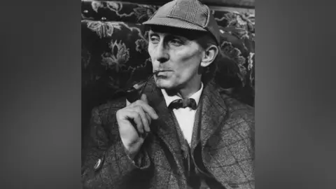 A black and white photo of a man in a cap and tweed jacket. He is smoking a pipe.