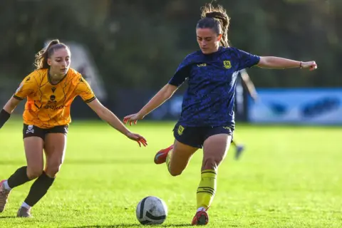 Lucy Copsey Photography Two female football players. One is in blue and is about to strike the ball. A second player in yellow is chasing her down from behind