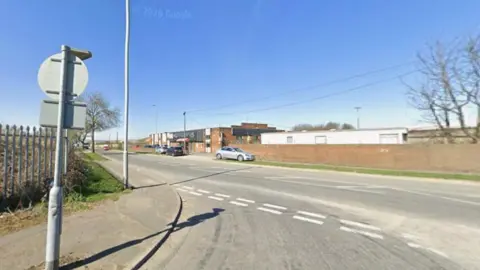 A wide road junction on a clear, sunny day. In the foreground, there is a pavement with a metal signpost and a tall security fence running alongside it. Several cars are driving along the road, which has white painted lane and junction markings.
On the right, a long brick wall lines the roadside, with low industrial buildings behind it.