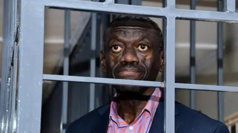 Reuters Kizza Besigye melihat melalui kisi-kisi logam di Pengadilan Militer Umum Uganda yang terletak di pinggiran Makindye di Kampala, Uganda 20 November 2024