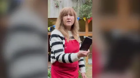 A woman outside with red apron holding a black phone.