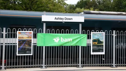 A sign saying Ashley Down on a railway platform above some railings. There is a train behind it. There are two GWR advertising posters pinned to the railings and a wide green banner that says 'bam' in white writing.