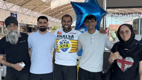 Five people pose beside displays at the Striving for Unity exhibition in The Galleria, Hatfield. South Asian Heritage Month banners, academy memorabilia and blue star-shaped balloons are visible around the exhibition stand.