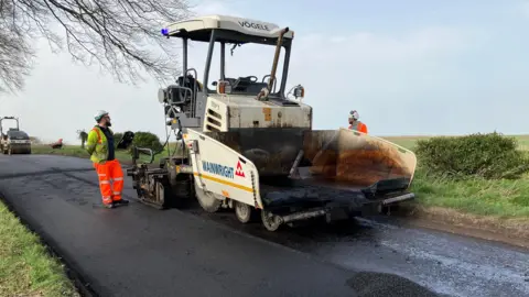 A large machine called a paver is spreading fresh tarmac onto the prepared road surface making it look like new