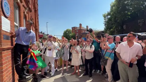 BBC A crowd gathers outside a red-brick building as a man stands on a small ladder pointing at a plaque. A rainbow flag is visible near the front. It’s a sunny day with trees and buildings in the background.