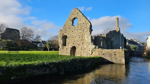 The remains of the medieval Christchurch Castle and Norman House stand on the banks of a river surrounded by grass. More modern development can be seen in the background and the sky is a vivid blue in bright sunshine.