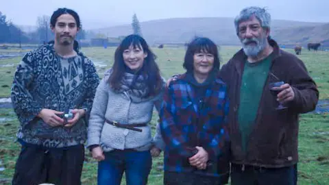 Holmes Family A family picture with, from left to right, a young man with long, dark hair pulled back in a patterned jumper, a woman with long, dark hair in a grey jacket with a black belt around her waist, a woman with shorter dark hair and a blue and black coat and a man with grey hair and a beard in a dark jacket holding a drink.