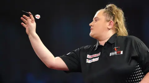 PA Wire Profile shot of Beau Greaves throwing a dart. She is wearing a short-sleeved, black sports shirt patterned with white spots on the sides