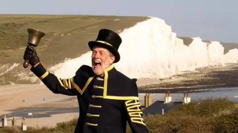 A man in a dark blue town crier uniform with gold trim and buttons, and top hat. He is holding a small bell on a handle and appears to be shouting. White cliffs and a beach can be seen in the background.
