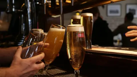 PA Media A bartender's hand holding glasses being filled with golden coloured larger from taps behind a bar with customers in the background. 