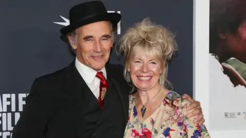 Getty Images Claire Van Kampen, who has a fair complexion and wears a floral dress with large silver earrings and a pendant necklace, smiles when her husband Mark Rylance puts his hand on her shoulder during a movie screening Are. 