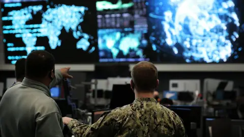 Two us cyber command officers in front of giant screens with data and information on them