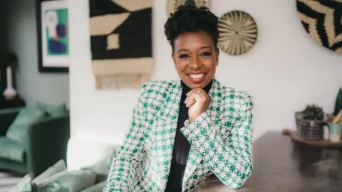 PA Media A headshot of saxophonist YolanDa Brown wearing a white and green checked suit jacket and black collared shirt. YolanDa is sitting at a table, resting her chin on her hand and smiling. In the background there are black and white artworks hanging on the wall.