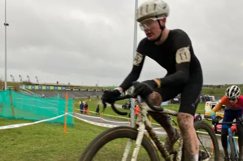 A rider in a black jersey is coated in mud up his bare legs, chest and on his visor and helmet. A rider in red is close behind him.