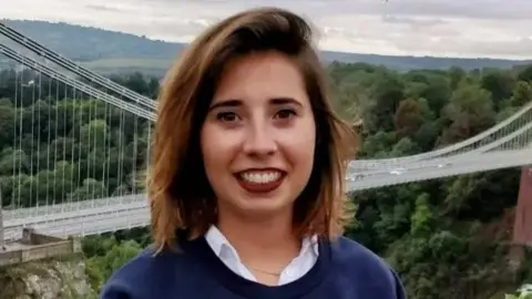 Marta Elena Vento smiles at the camera. Clifton Suspension Bridge is in the background. She has shoulder-length brown hair and wears a white shirt, blue top and make-up.