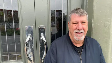 Carl Chinn, a man with silver hair and a beard, smiles at the camera while standing in front of a pair of green doors.