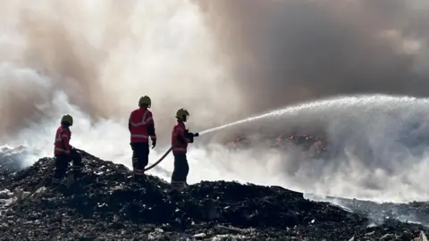 Three firefighters are wearing yellow helmets, red jackets and black trousers. They are standing on a pile of burnt rubbish on the left. One of them is holding a hose pipe trying to put out a fire.