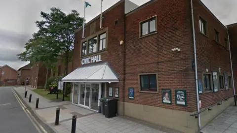 The former Stanley Civic Hall. The brown brick building has glass dorrs at its entrance. There are two flag poles attached to the roof above the entrance. Posters are displayed in frames around the building.