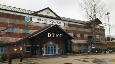 A general view picture of The Avenue Stadium in Dorchester, which is a brick building, with an entrance of two blue doors with DTFC above them.