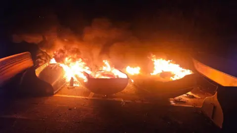 A row of boats are engulfed by large orange flames. It is dark. Smoke is rising into the sky.