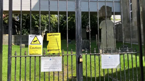 A gate made of black metal railings is closed and padlocked. There are signs explaining why the burial ground is closed. Behind the gate is an area of green grass bathed in sunlight and gravestones in the shadow of a nearby building.