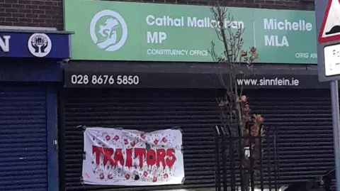 BBC A wide shot of the office of Michelle O'Neill and Cathal Mallaghan. A green sign bears their names and the Sinn Féin party logo. Below it are black shutters, which a banner is stuck to. It reads "Traitors" in red writing made to look like blood and with bloody hand prints. The "O" is styled as a Remembrance poppy.