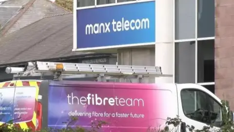 A van with colourful Manx Telecom branding on its rear and driver side panel and a ladder on its roof. Its parked outside a building with blue and white sign that reads Manx telecom.