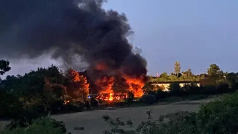 The skip lorry site in College Road, Epsom on fire, with thick black smoke and flames filling the air.