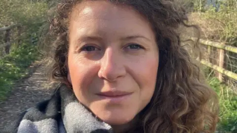 Jess Laurie A close up of a woman's face, wearing a black and grey scarf. A country lane with wooden faces can be seen in the background.