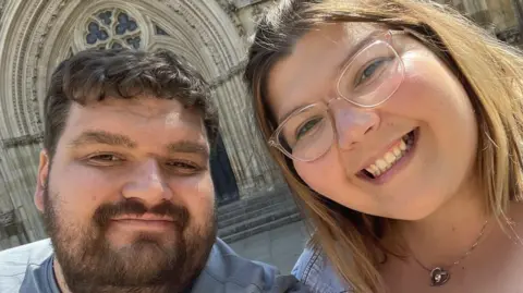 Tom Brakewell and Gabby Evans are smiling in a selfie. Tom has brown hair and a beard and Gabby has shoulder length blonde hair.