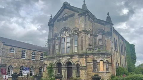 The picture is a large, historic church building constructed from yellowish-brown stone, featuring classical and Gothic revival architectural elements. The structure has a tall, rectangular form with a prominent central façade facing the street. The sky above is filled with dark, heavy clouds, giving the scene a slightly gloomy, dramatic atmosphere.