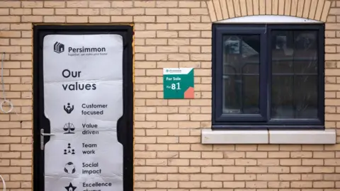 A For Sale sign for an apartment at a Persimmon Plc residential property construction site, with tan bricks and a sign on the door listing Persimmon's values as a housebuilder, including being customer focused, value driven and social impact. 