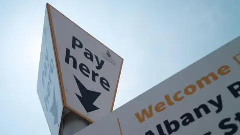 A close-up picture looking up at a Cornwall Council car park sign taken on a clear sunny day. The signs says 'Pay here' in bold black text. 