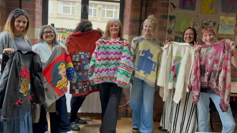 BBC/Aisha Iqbal Seven women stand in a line holding up jackets and jumpers they have made from recycled materials