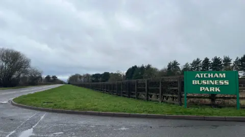 ELLEN KNIGHT/BBC Photograph of the turning into Atcham Business Park. On the right side of the photo, a large sign is staked into the ground, reading 'Atcham Business Park' in light yellow text on a dark green background. On the left, the B-road stretches away into the distance, with the smaller access road running across the bottom of the photograph. A green grass verge runs either side of the main road, with fencing and a brown hedgerow on the right. In the distance, a row of coniferous trees can be seen lining the horizon. The sky is grey and overcast, and there are puddles on both stretches of road indicating recent rain. 