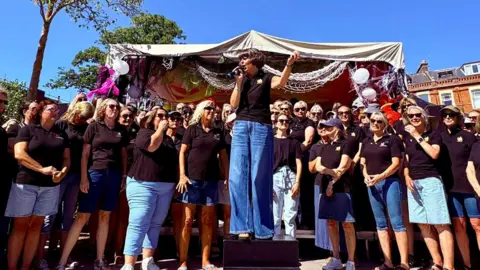 Alex Walton Photography A big group of women wearing black tshirts and denim jeans or skirts. Sam is standing on a box and speaking into a microphone with one extended arm. A gazebo is behind them strung with nets and balloons