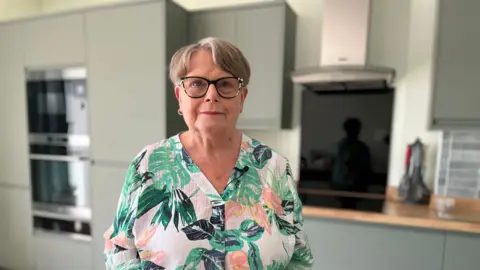 Carole Hager stands in the kitchen of her newly rebuilt house. She has short grey hair and is wearing black-rimmed glasses and a brightly coloured floral shirt. Behind her is a modern kitchen with sage green kitchen cupboards and wooden work surfaces. 