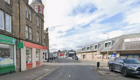 A view down Tomnahurich Street in Inverness. The street is lined with traditional-style buildings on one side with small shops on the ground floor, and across the road is a Tesco supermarket.