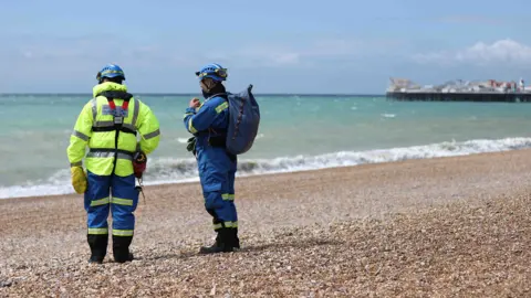 Two responders in reflective gear stand on the pebble beach of Brighton looking out to sea with the Brighton pier in the background on Wednesday.