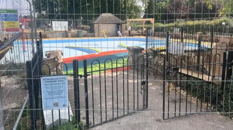Barriers around an empty paddling pool in a park. A sign says St Nicholas Park Paddling Pool. A skip and cardboard boxes can be seen around the outside of it.
