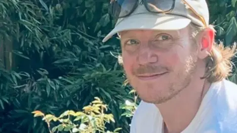 JustGiving James also known as Jamie Boland with stubble wearing a beige baseball cap with a pair of sunglasses on top in front of foliage smiling